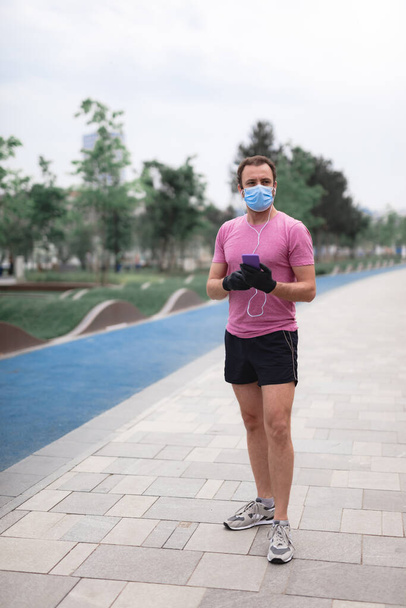 Esportista com máscara médica e luvas, smartphone e fones de ouvido trabalhando, correndo em ambientes urbanos. - Foto, Imagem