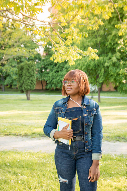 La chica pensativa mira a la distancia. Joven chica negra hermosa de pie en el parque sosteniendo un cuaderno en sus manos. Retrato de una estudiante al aire libre
. - Foto, imagen