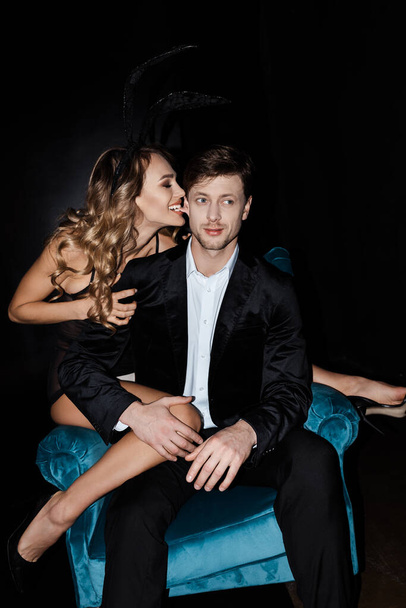 Beautiful woman biting ear of handsome boyfriend in suit sitting in armchair isolated on black  - Фото, изображение