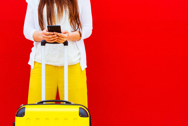 woman with a yellow suitcase reading messages on her mobile phone leaning against a red wall - Foto, Imagem