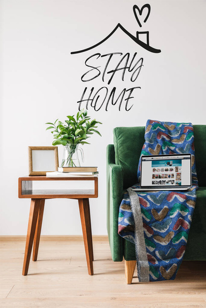 KYIV, UKRAINE - APRIL 14, 2020: stay home lettering near green sofa, blanket, laptop with amazon website and wooden coffee table with green plant - Photo, image