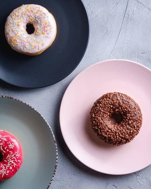 Three donuts on plates, chocolate, pink and vanilla donut with sprinkles, sweet glazed dessert food on concrete textured background, top view - Fotografie, Obrázek