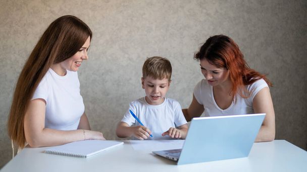 Two women help the boy do school homework. Female tutor teaches a child at home in the kitchen. The school teacher conducts an individual lesson. Mother helps her son to study. - Фото, изображение