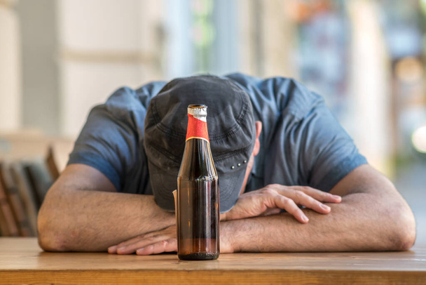 Pensieve en trieste man met gebogen hoofd zittend met een fles bier buiten een bar. Selectieve focus - Foto, afbeelding