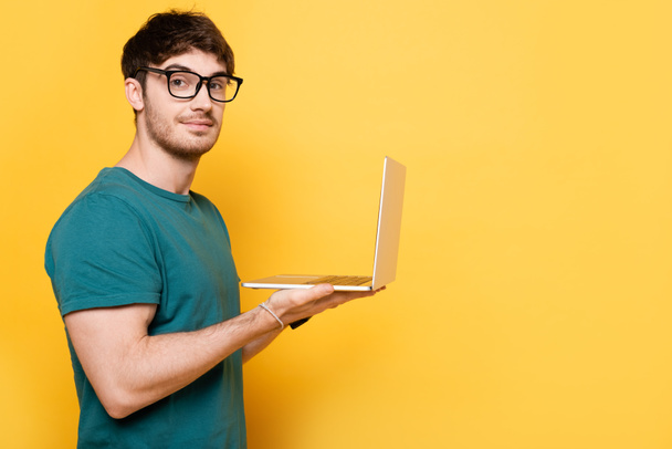 smiling young man looking at camera while holding laptop on yellow - Φωτογραφία, εικόνα