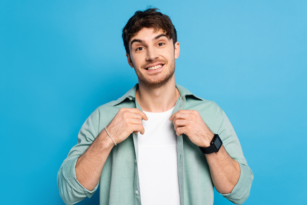handsome young man touching shirt while smiling at camera on blue - Foto, imagen
