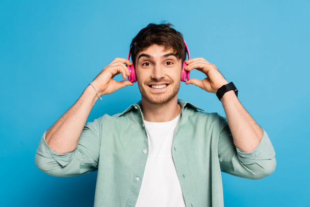 szczęśliwy młody człowiek dotykając bezprzewodowych słuchawek podczas słuchania muzyki na niebiesko - Zdjęcie, obraz