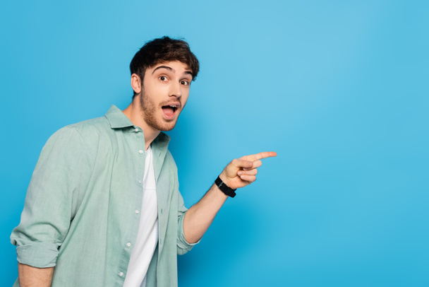 shocked young man looking at camera and pointing with finger on blue - Фото, зображення