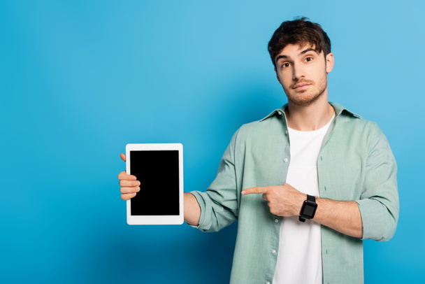 όμορφος νεαρός άνδρας με το δάχτυλο στο ψηφιακό tablet με λευκή οθόνη στο μπλε - Φωτογραφία, εικόνα