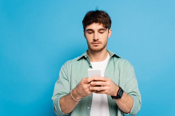 serious young man chatting on smartphone isolated on blue - Photo, Image