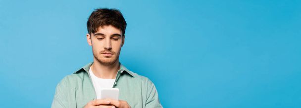 horizontal image of serious young man using smartphone isolated on blue - Photo, Image