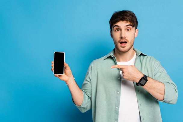 jovem surpreso apontando para o smartphone com tela em branco no azul
 - Foto, Imagem