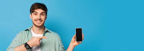 website header of happy young man pointing at smartphone with blank screen on blue - Photo, Image