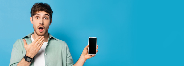 horizontal image of shocked young man showing smartphone with blank screen on blue - Photo, Image