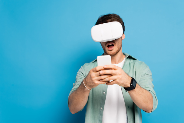 shocked young man in vr headset using smartphone on blue - 写真・画像