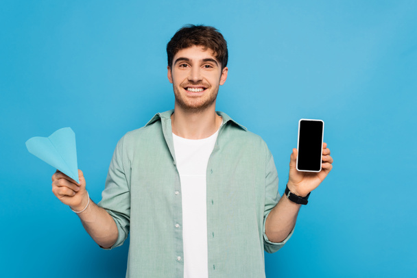 gelukkig jonge man tonen smartphone met leeg scherm en papier vliegtuig op blauw - Foto, afbeelding