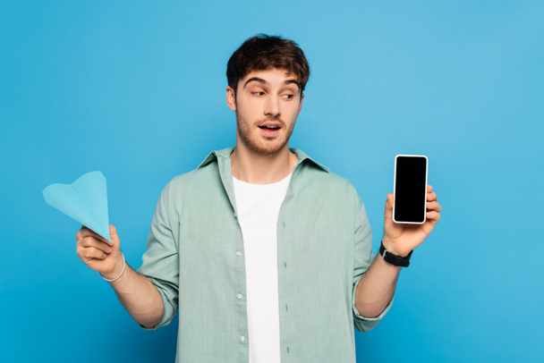 přemýšlivý mladý muž drží smartphone s prázdnou obrazovkou a papírové letadlo na modré - Fotografie, Obrázek