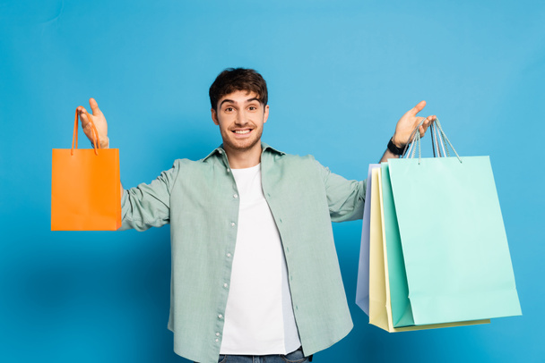 happy young man holding colorful paper bags while looking at camera on blue - Photo, Image