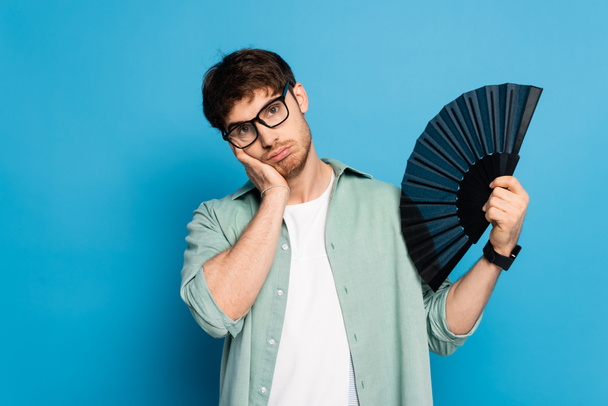 exhausted young man touching face while holding hand fan on blue - Foto, imagen