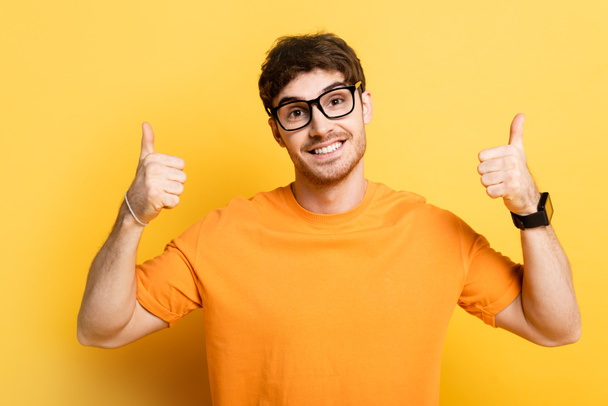 cheerful young man smiling at camera and showing thumbs up on yellow - Фото, изображение