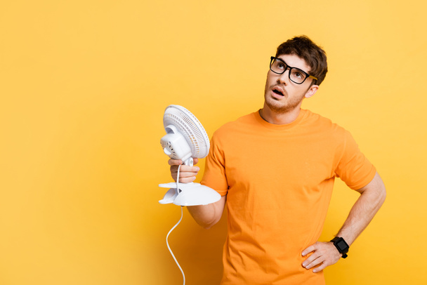 nespokojen mladý muž vzhlíží, zatímco trpí horkem a drží elektrický ventilátor na žluté - Fotografie, Obrázek