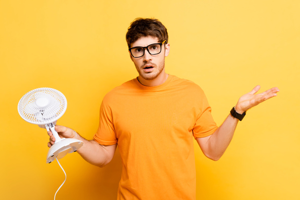 discouraged man holding electric fan and showing shrug gesture on yellow - Foto, imagen