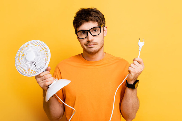 disappointed young man holding electric fan while looking at camera on yellow - Фото, изображение