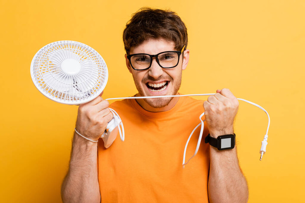 angry man in eyeglasses holding broken electric fan on yellow   - Fotografie, Obrázek