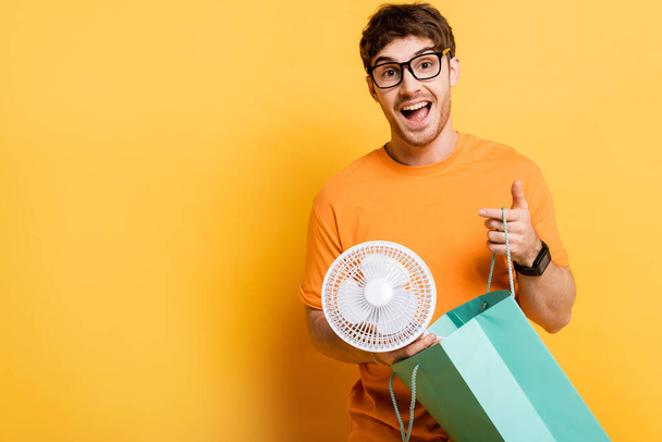 surprised young man in eyeglasses holding shopping bag with new electric fan on yellow   - Fotoğraf, Görsel