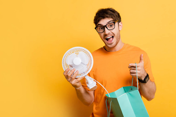 excited man in eyeglasses holding shopping bag with new electric fan on yellow   - Foto, Imagem
