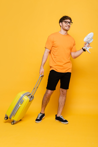 glimlachende man met reistas met elektrische ventilator op geel  - Foto, afbeelding