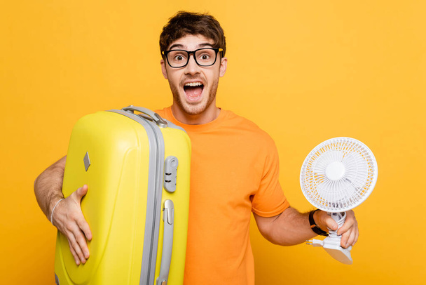 excité homme tenant sac de voyage et ventilateur électrique sur jaune  - Photo, image