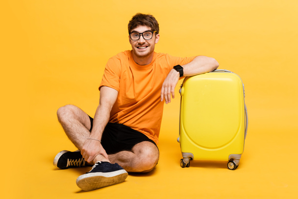 joyeux touriste masculin assis près du sac de voyage pour les vacances d'été sur jaune  - Photo, image