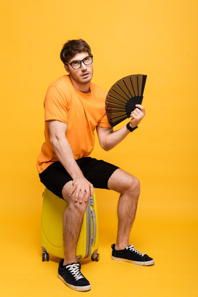 frustrated man suffering from heat while using hand fan and sitting on suitcase on yellow  - Foto, Bild