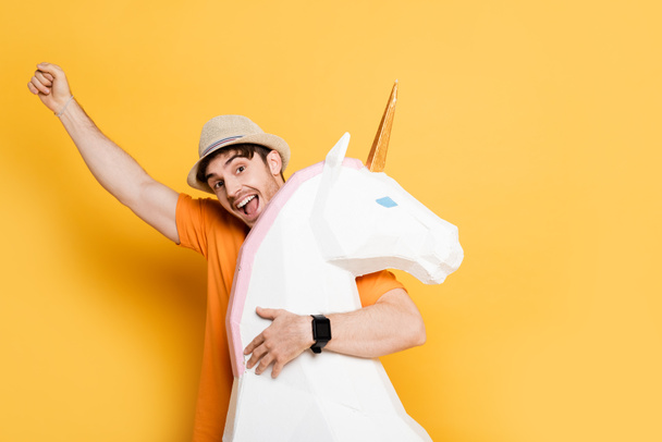 excited man in summer hat holding fist up and hugging decorative unicorn on yellow  - Photo, Image