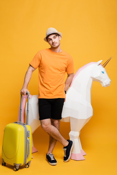 νεαρός άνδρας ταξιδιώτης με καπέλο στέκεται με μονόκερο και βαλίτσα στο κίτρινο - Φωτογραφία, εικόνα