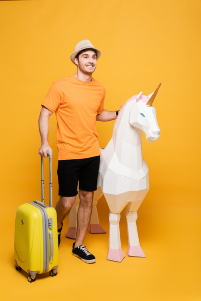 smiling traveler in hat standing with unicorn and suitcase on yellow - Foto, immagini