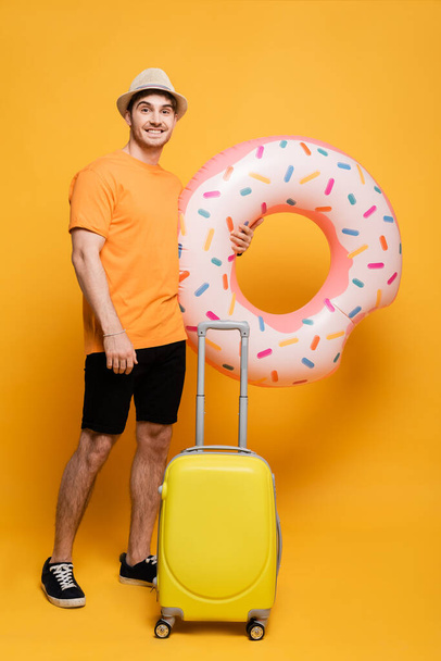 happy man in hat with suitcase and inflatable donut ready for summer vacation on yellow - Photo, Image