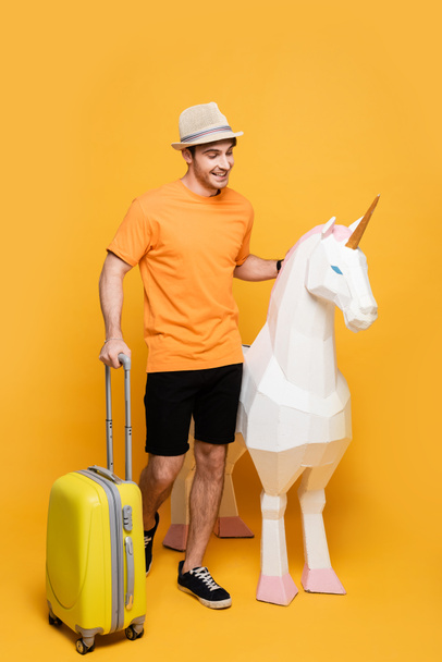 cheerful traveler in hat standing with unicorn and suitcase on yellow - Фото, зображення