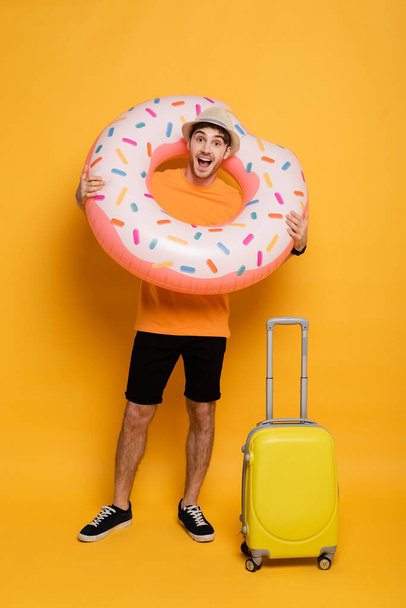 opgewonden man met koffer en opblaasbare donut klaar voor zomervakantie op geel - Foto, afbeelding