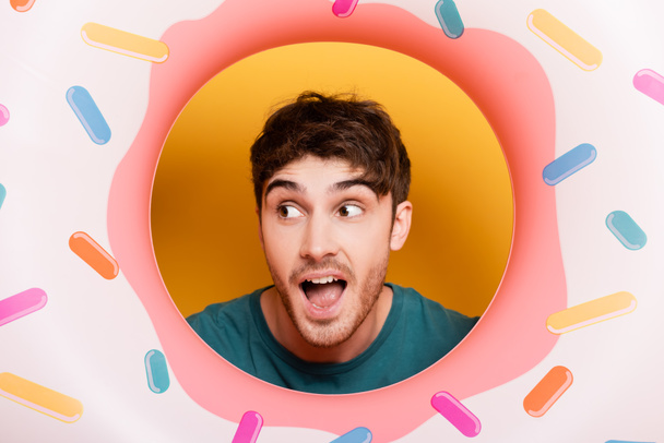surprised man with open mouth standing with inflatable donut on yellow  - Photo, Image