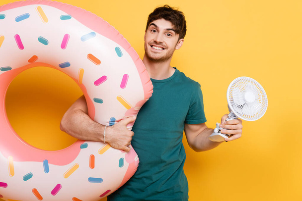 smiling man holding inflatable donut and little electric fan on yellow  - Фото, изображение
