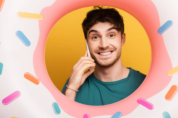 vrolijk jongeman met opblaasbare donut praten op smartphone op geel - Foto, afbeelding