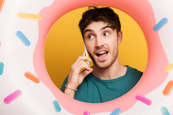 Aufgeregter Mann mit aufblasbarem Donut redet auf Smartphone auf gelb - Foto, Bild