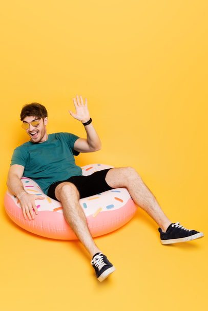 excited man in sunglasses waving hand and sitting in inflatable donut on yellow  - Photo, Image