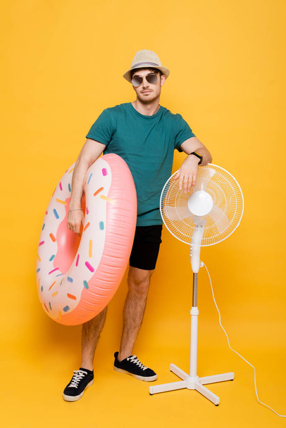 handsome man in sunglasses and hat holding inflatable donut while standing near electric fan on yellow  - Fotografie, Obrázek