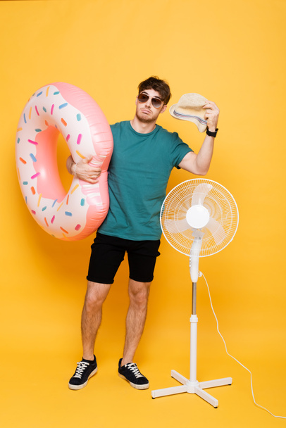 Verwirrter Mann mit Sonnenbrille hält Hut und aufblasbaren Donut in der Nähe von Elektroventilator auf gelb  - Foto, Bild