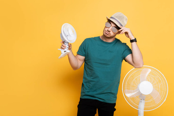 молодий чоловік в сонцезахисних окулярах і капелюсі стоїть з двома електричними вентиляторами на жовтому
    - Фото, зображення