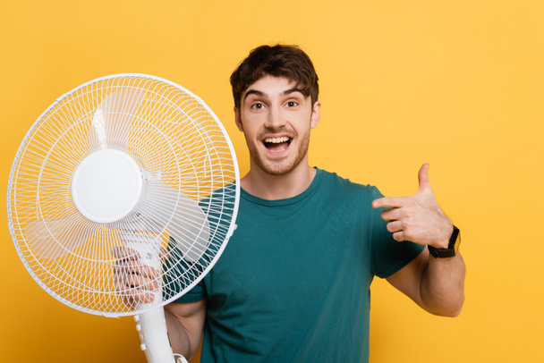 щасливий молодий чоловік вказує пальцем на електричний вентилятор на жовтому
     - Фото, зображення