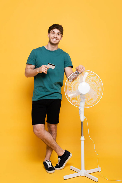 smiling young man holding credit card while standing with electric fan on yellow      - Foto, Bild
